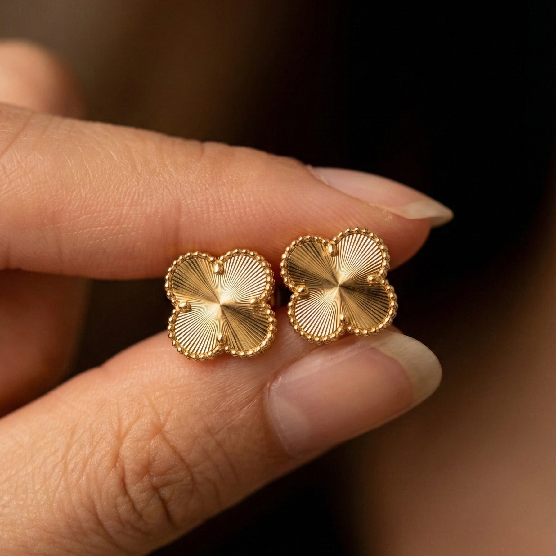 Eva Studs gold four leaf clover stud earrings with sunburst engraved petals and beaded gold trim held between fingers – tarnish-free waterproof stainless steel earrings for sensitive ears