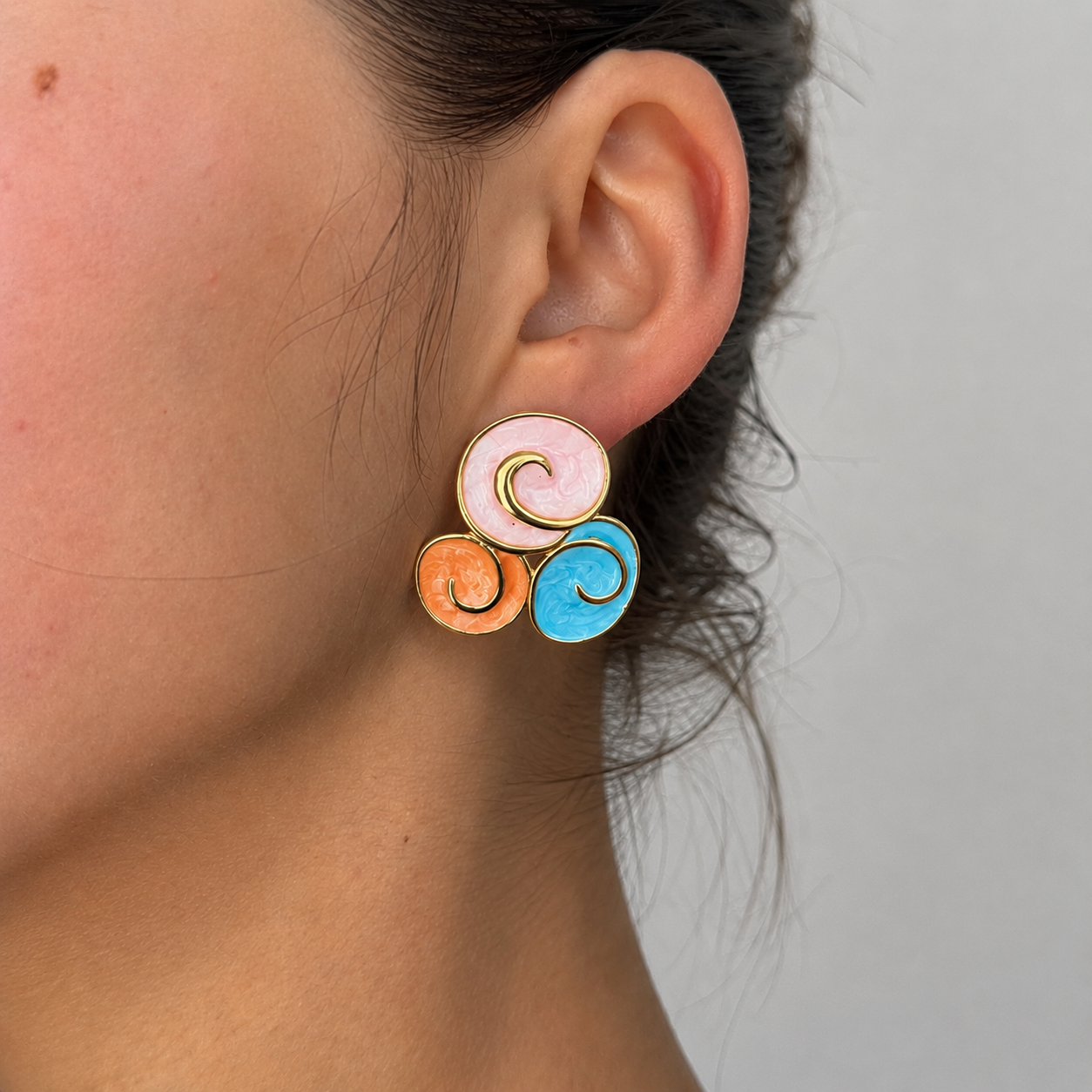 Kima Studs colourful enamel swirl stud earrings in pink, coral and blue with gold spiral detail on dark textured background – hypoallergenic statement stud earrings, tarnish-free surgical steel