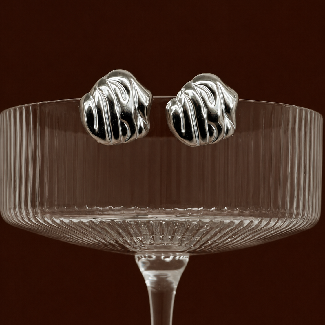Mmírí Studs chunky textured silver stud earrings with rippled molten wave surface balanced on ribbed cocktail glass against dark background – hypoallergenic tarnish-free sterling silver stud earrings